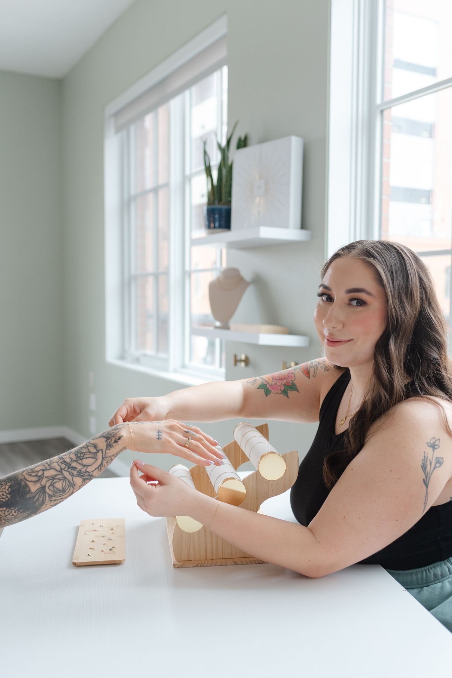 A person in a black tank top works at a white table near bright windows with visible tattoos and shelving on display.