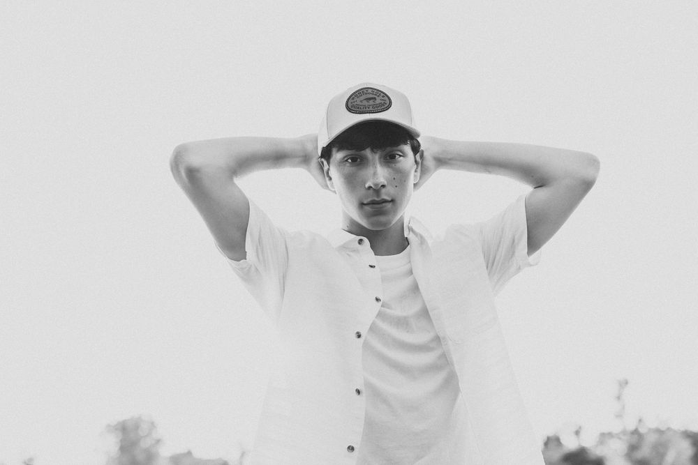 Young man in white shirt and cap poses with hands behind head in black and white outdoor photo.