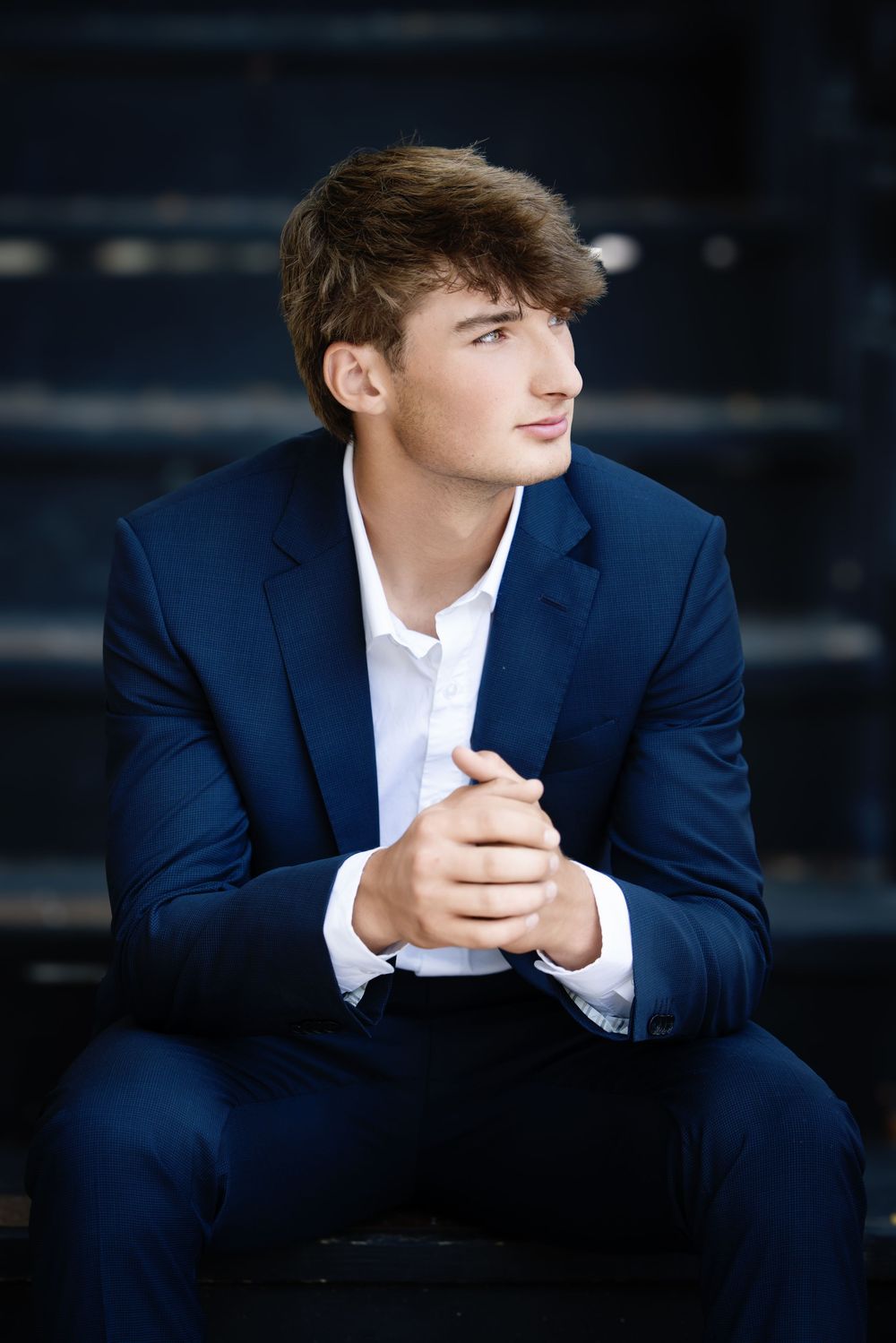 Young man in navy suit and white shirt poses thoughtfully on dark steps in professional portrait series.