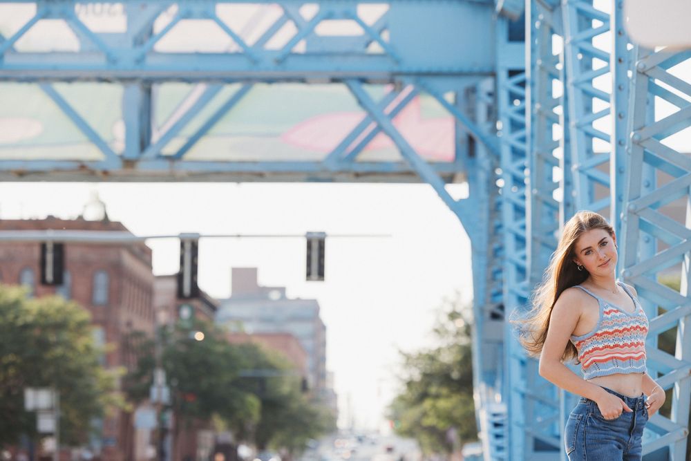 A person in a blue dress walks under a blue steel bridge structure on a sunny day.
