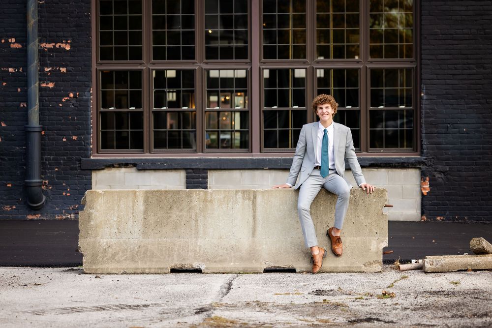 A person in a light blue suit jumps playfully across a concrete barrier in front of an industrial brick building.
