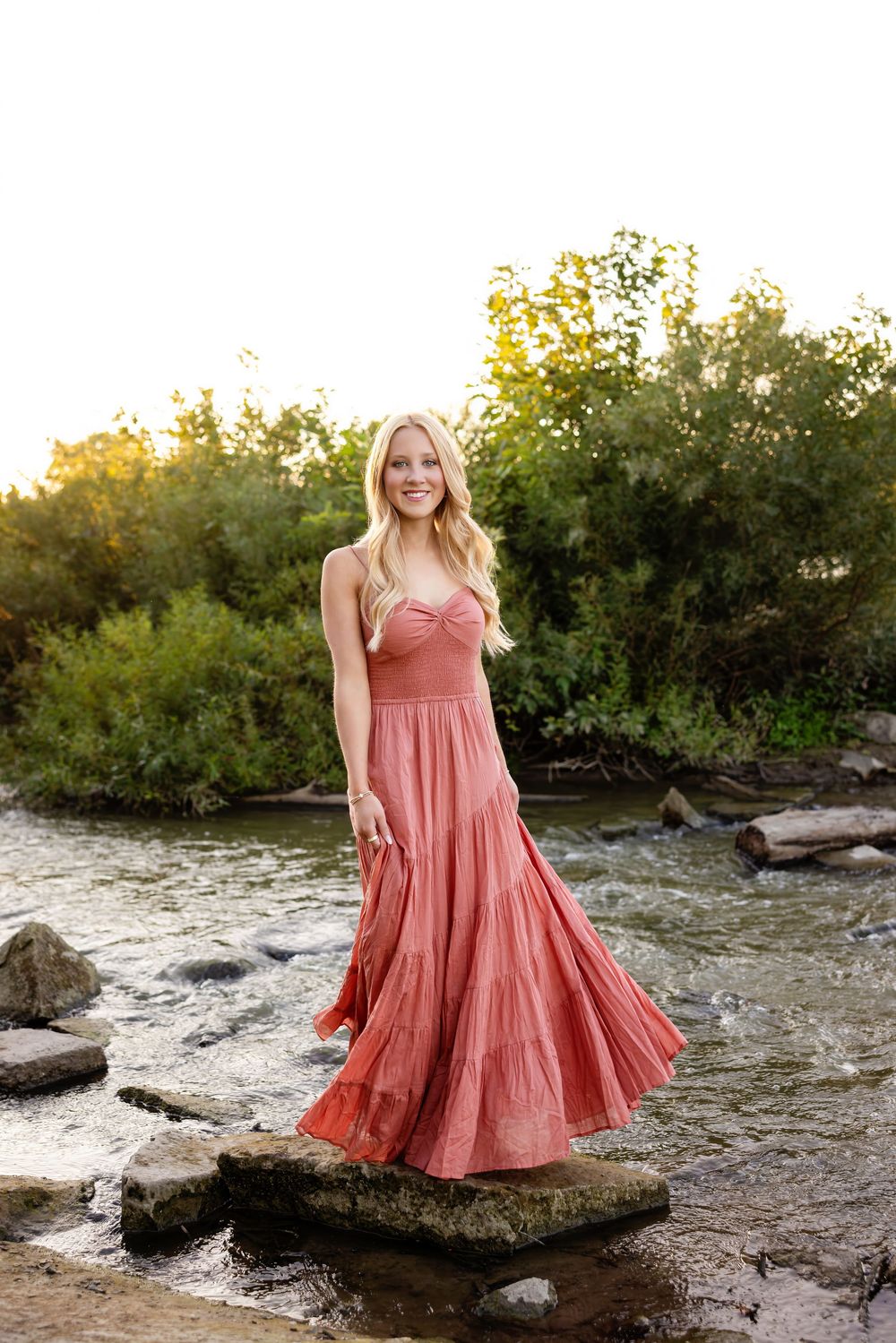 A person in a coral tiered dress poses gracefully on rocky terrain near water during golden hour.