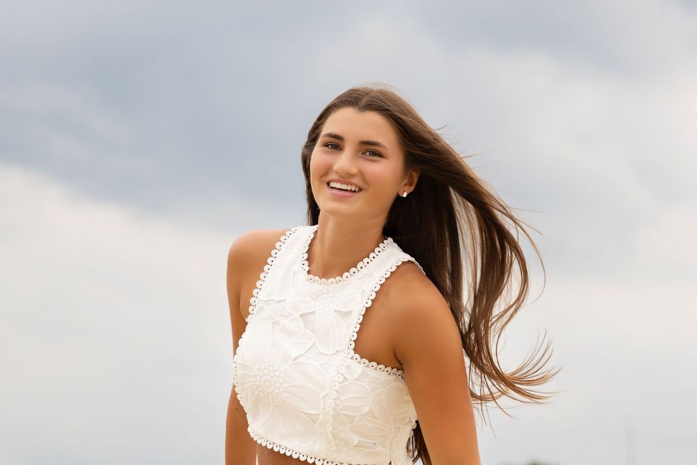A woman in white lace top poses outdoors with wind blowing through her hair against a cloudy sky.