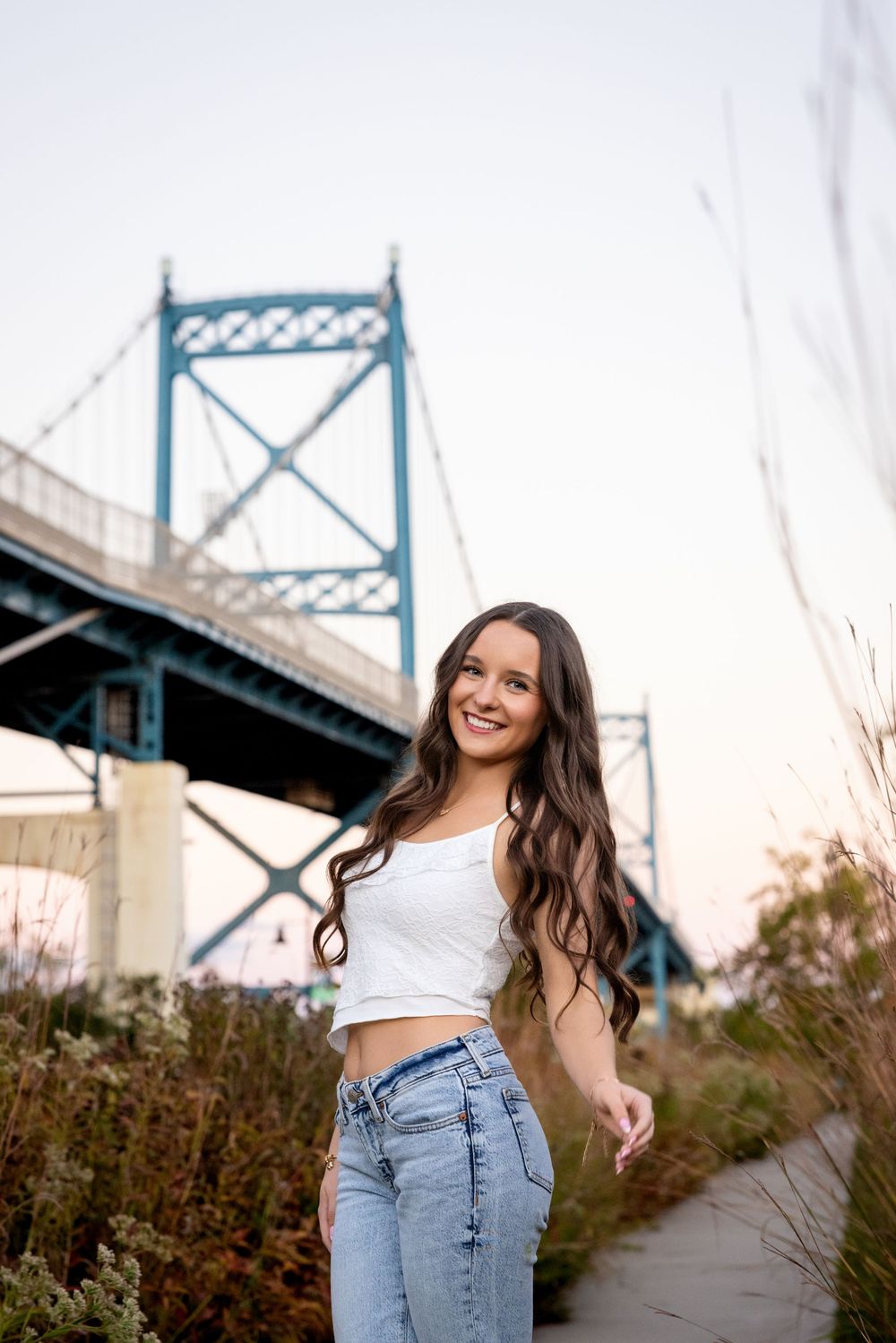 Young woman with long wavy hair in white crop top and jeans smiling outdoors near a large blue suspension bridge.