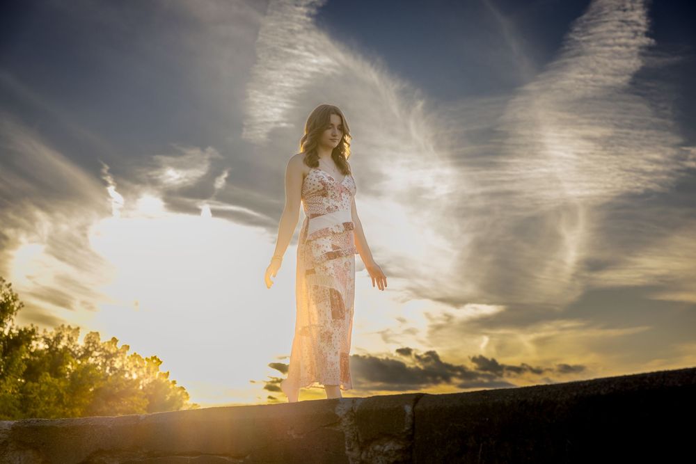 A silhouetted figure stands in a flowing dress against a dramatic sunset sky with rays of golden light streaming through clouds.