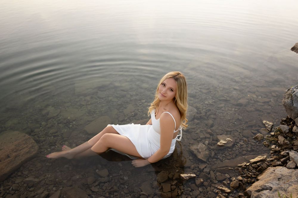Woman in white dress sitting at waters edge on rocky shore during sunset.