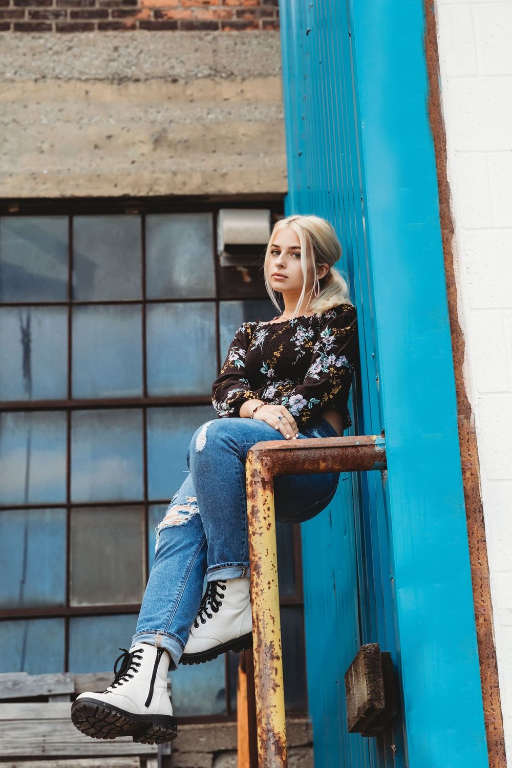 Young blonde woman in floral top, ripped jeans, and white boots sitting on rusty railing near blue wall.