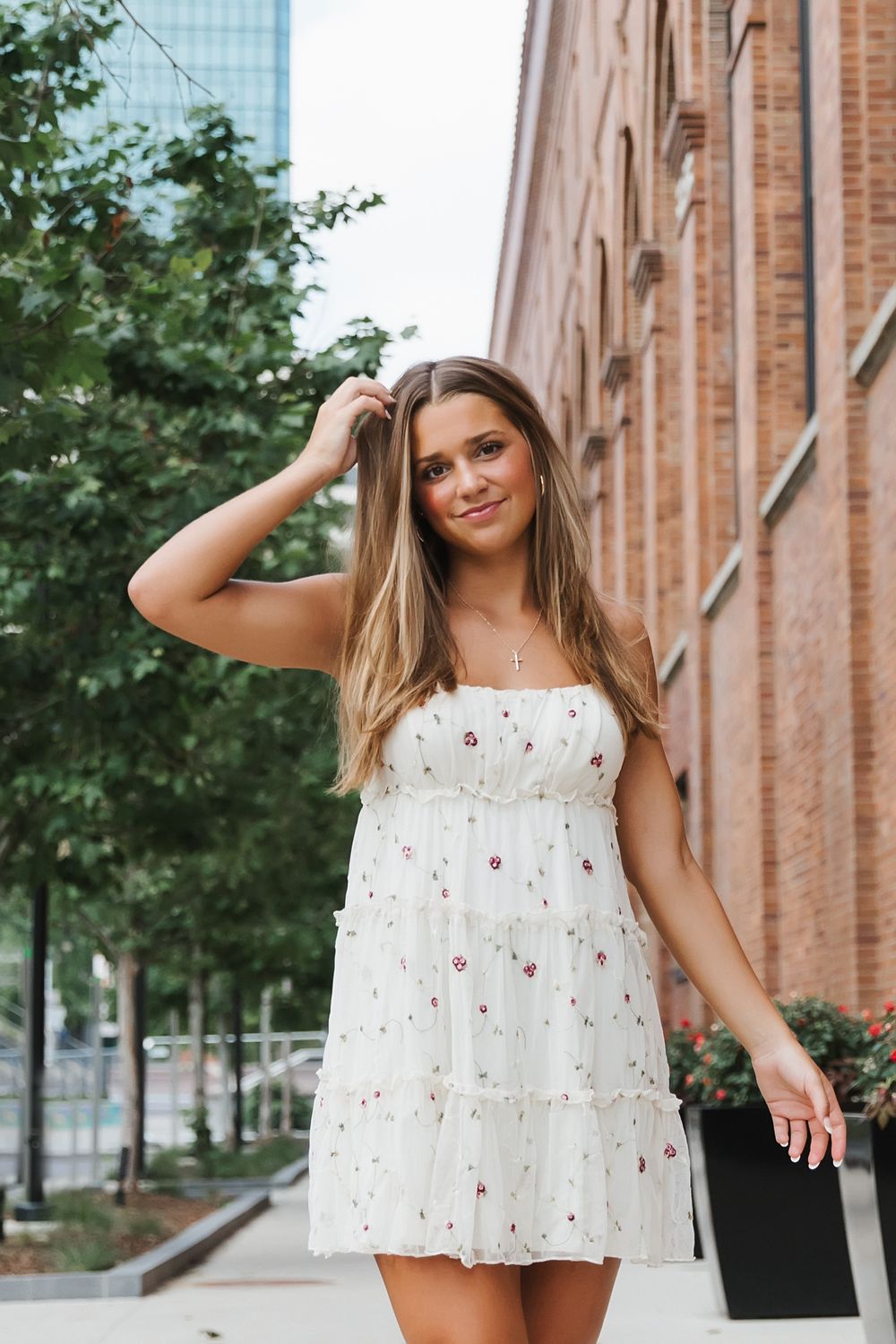 Young woman in white floral sundress poses outdoors near brick building and green trees on a city street.