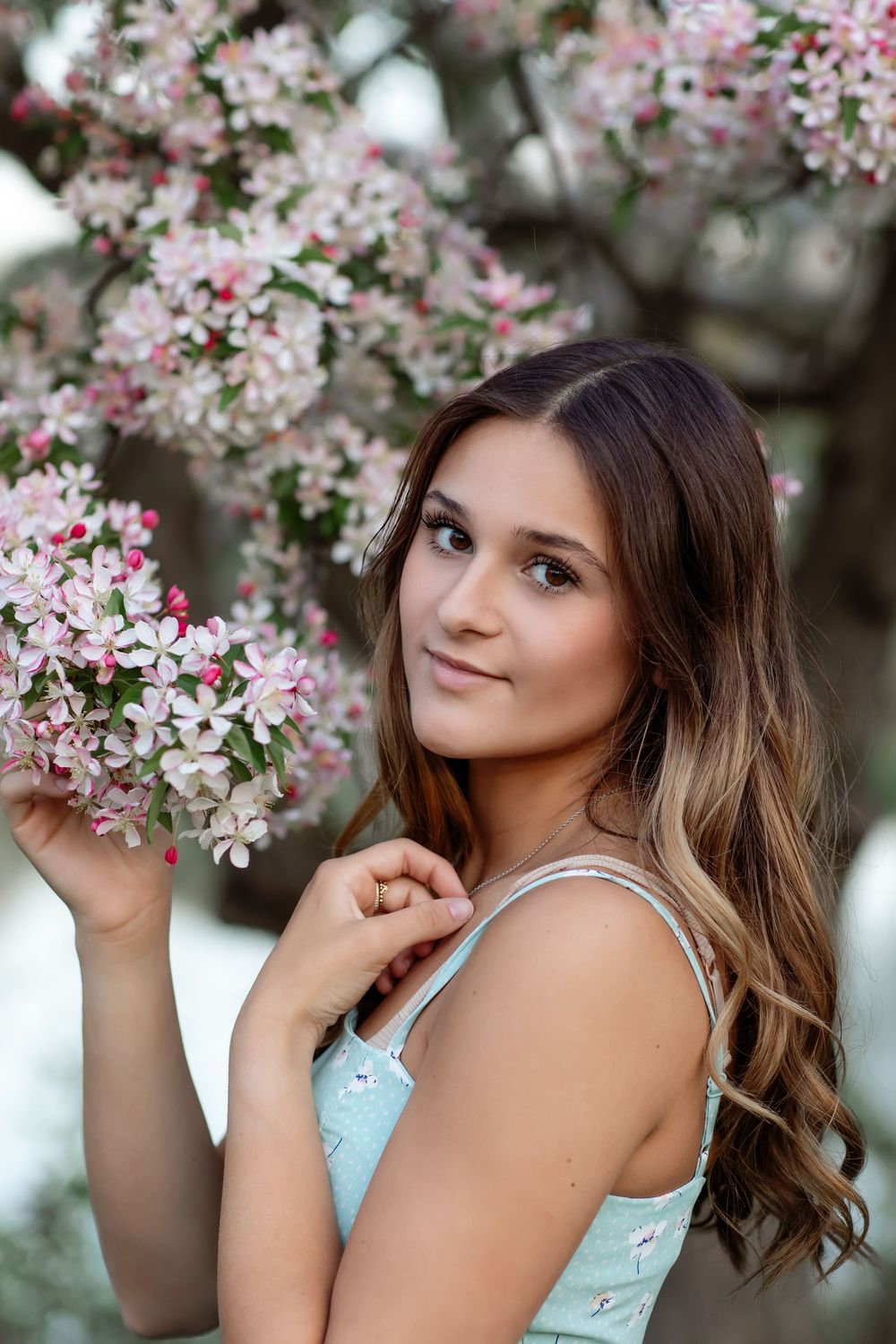 Young woman with long brown hair in a light blue dress poses near pink blossoming tree branches outdoors.