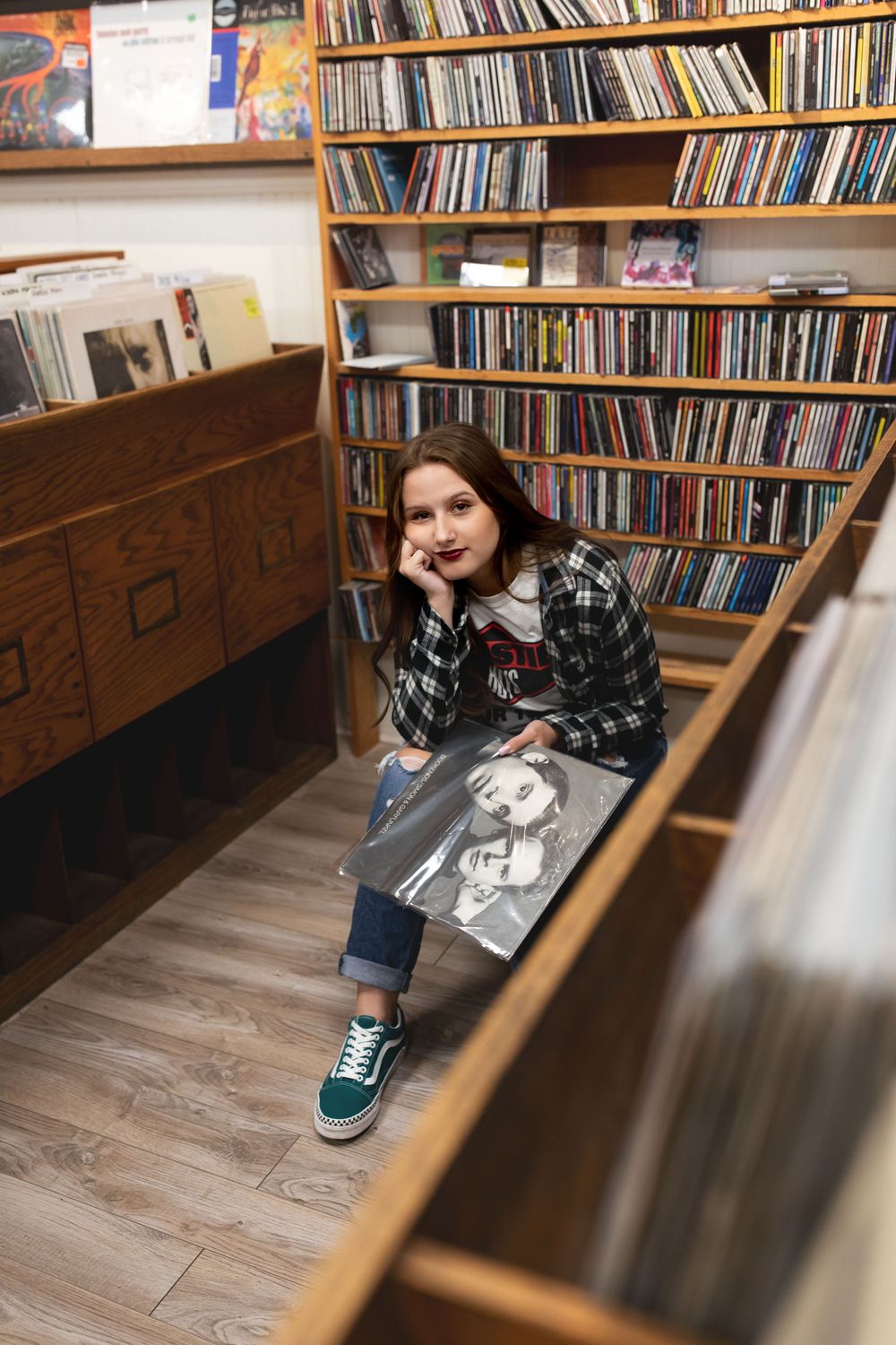 A person sits on the floor of a record store browsing vinyl albums, surrounded by wooden shelves filled with CDs and records.