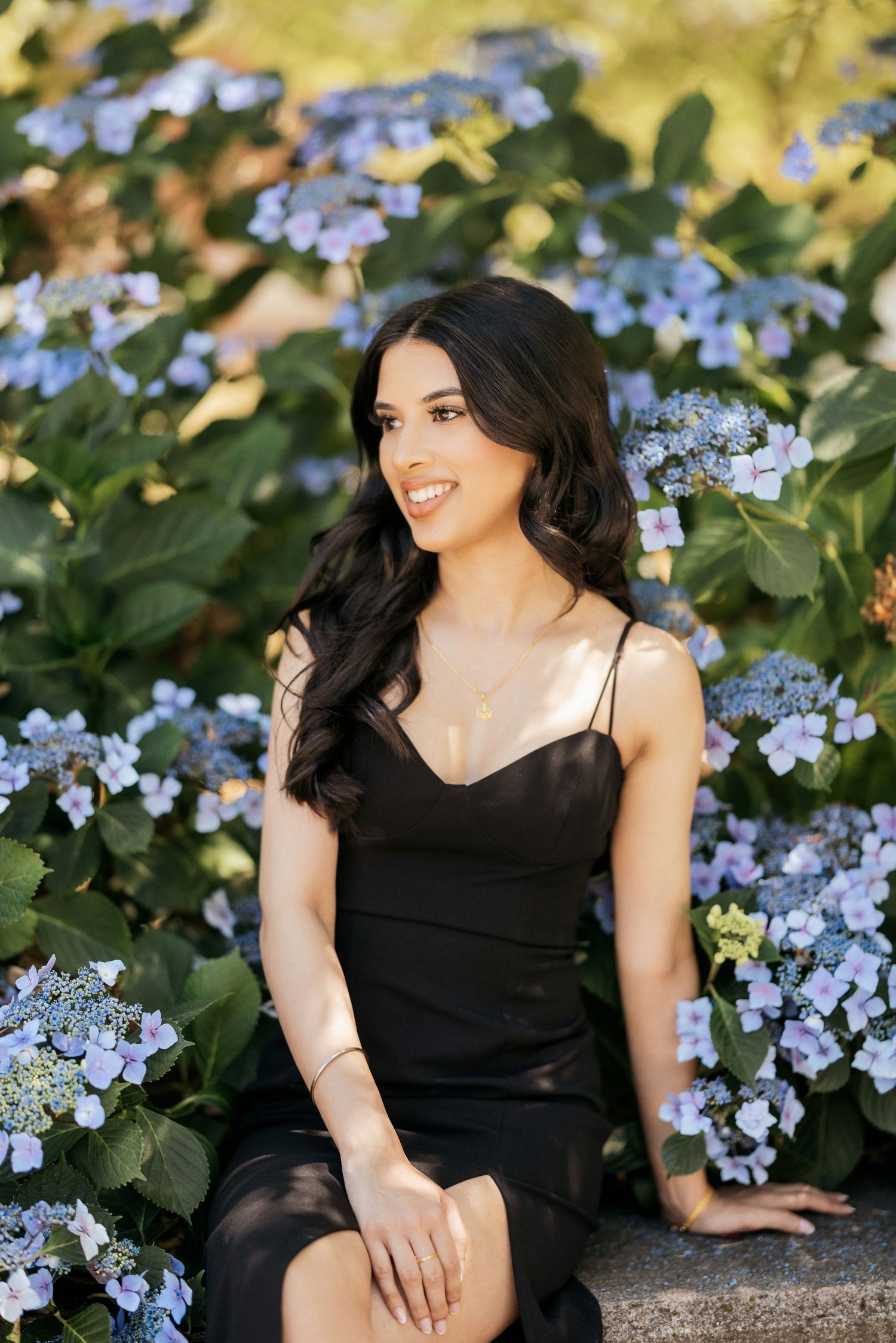 Graduate in black dress poses by blooming blue hydrangea bush.