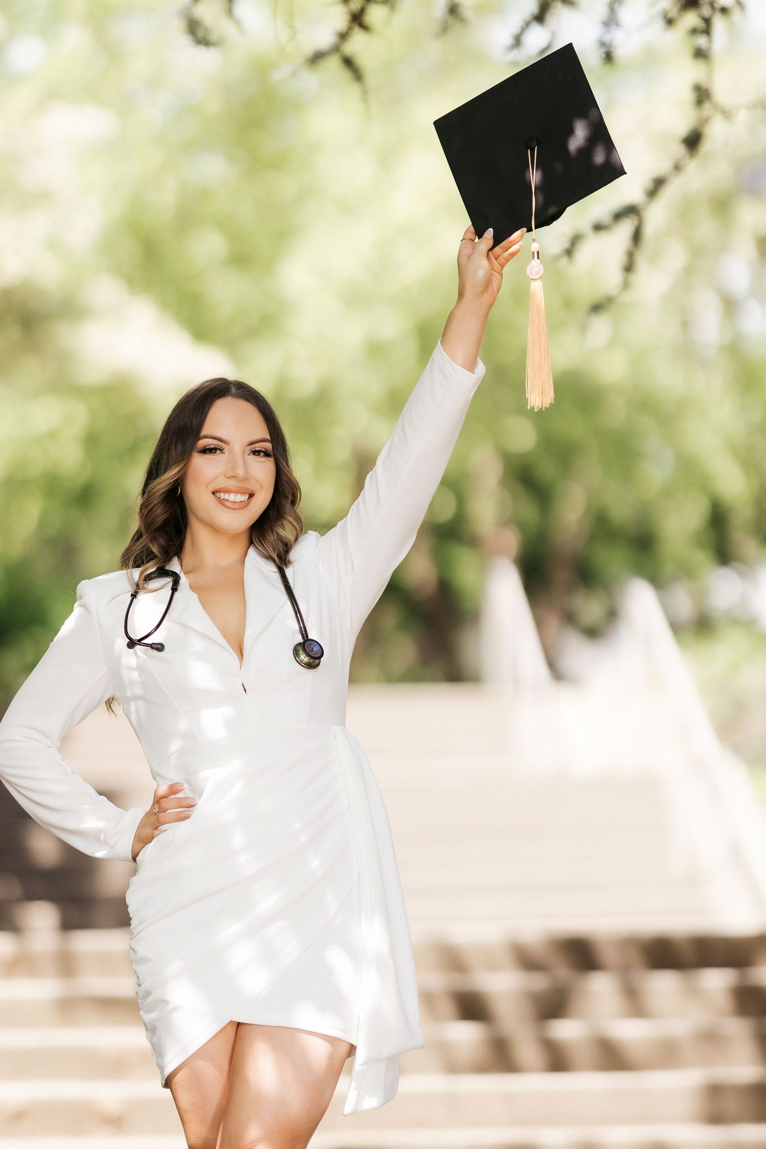 Medical graduate in white dress celebrating with raised cap outdoors.