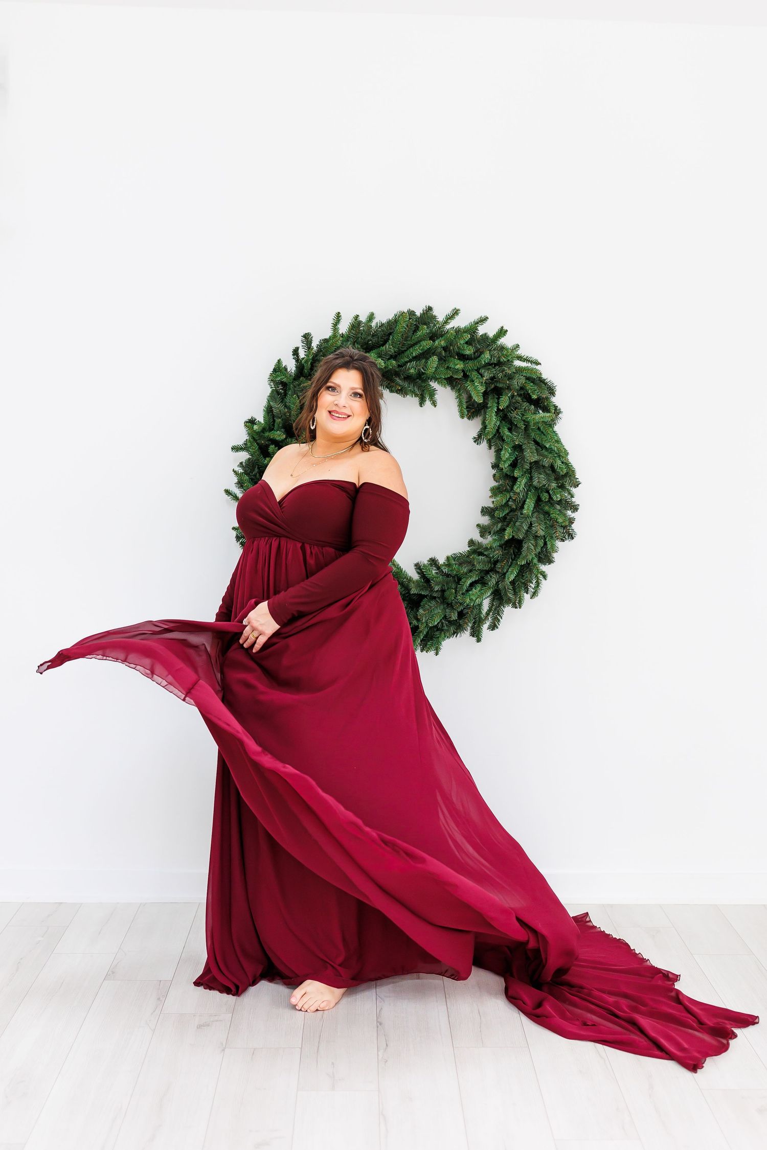 A series of artful maternity portraits featuring a burgundy flowing gown and evergreen wreath against a white backdrop.
