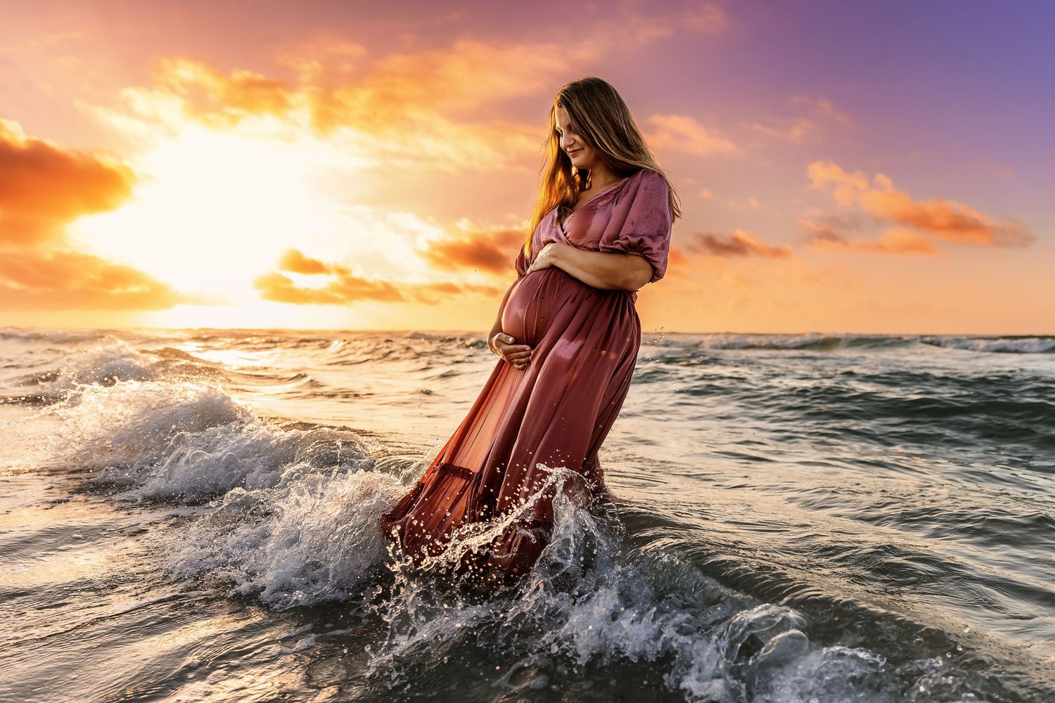 A person in a flowing red dress stands in ocean waves during a dramatic golden sunset at the beach.
