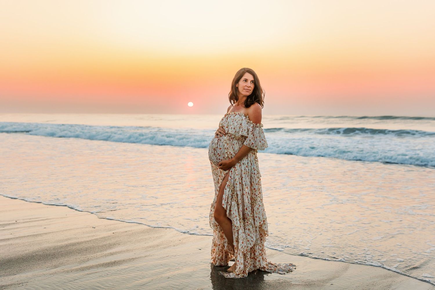 A pregnant person wearing a flowy dress poses on a beach at sunset with ocean waves and golden sky in the background.