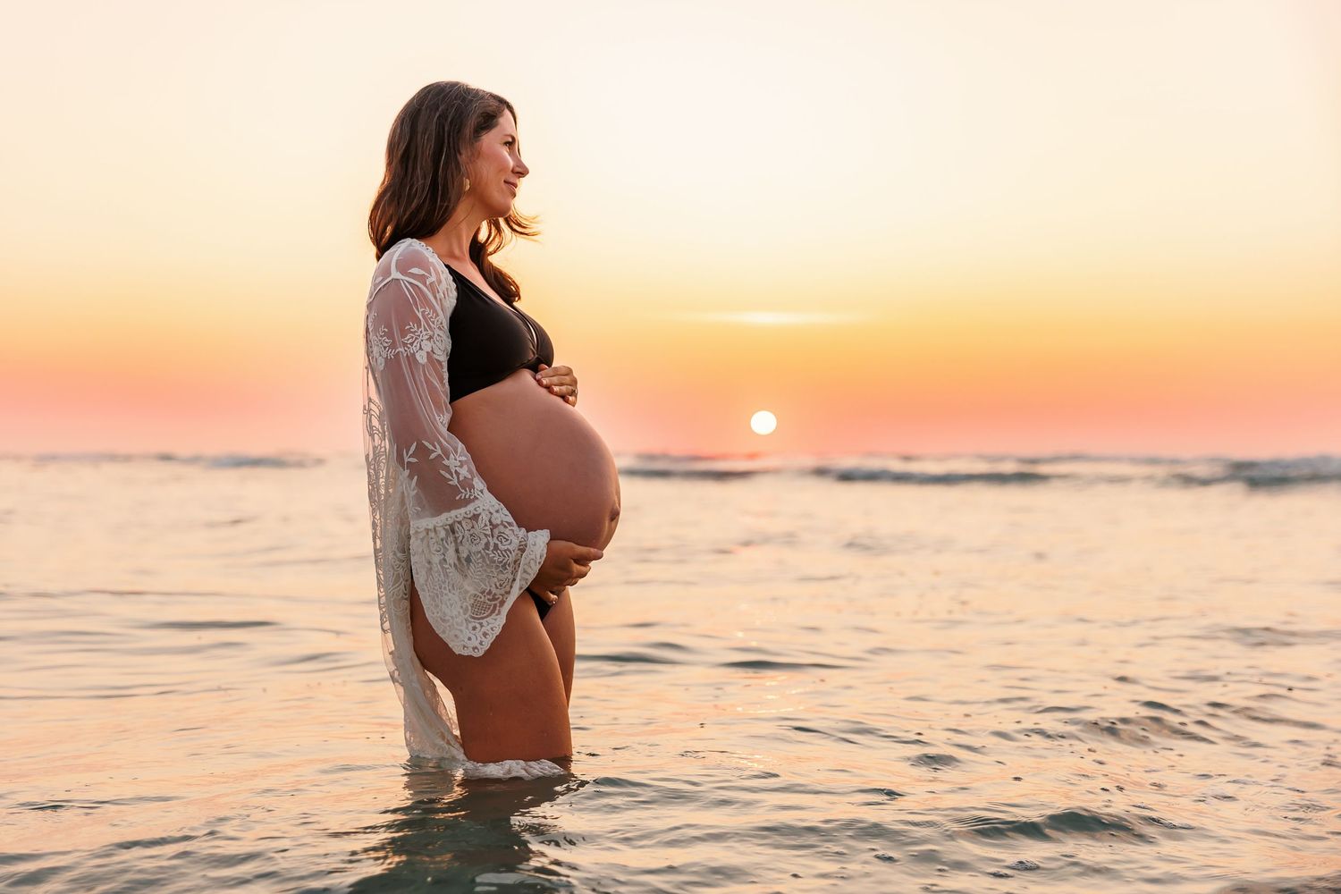 Expectant mother standing in ocean water at sunset wearing black bikini and white cover up.