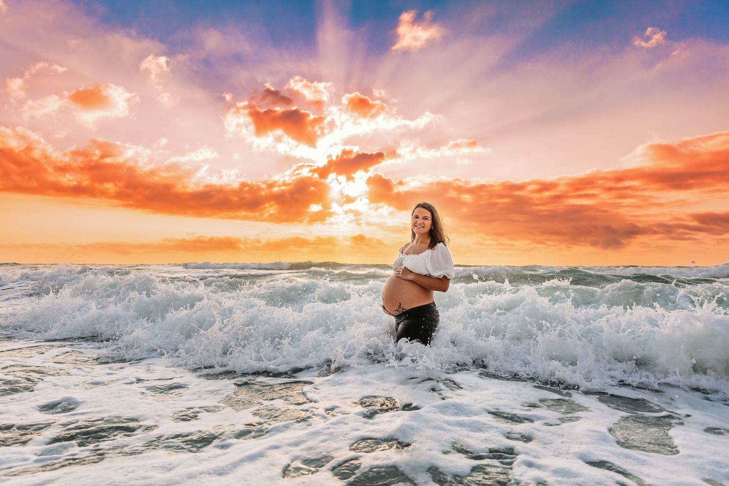 A person stands in crashing ocean waves at sunset with dramatic orange clouds in the sky above.