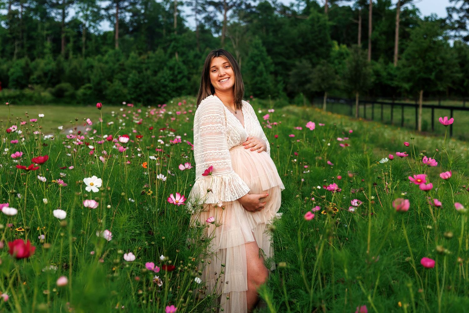 A field of pink poppies blooms alongside someone in a cream-colored dress on a lush green hillside.