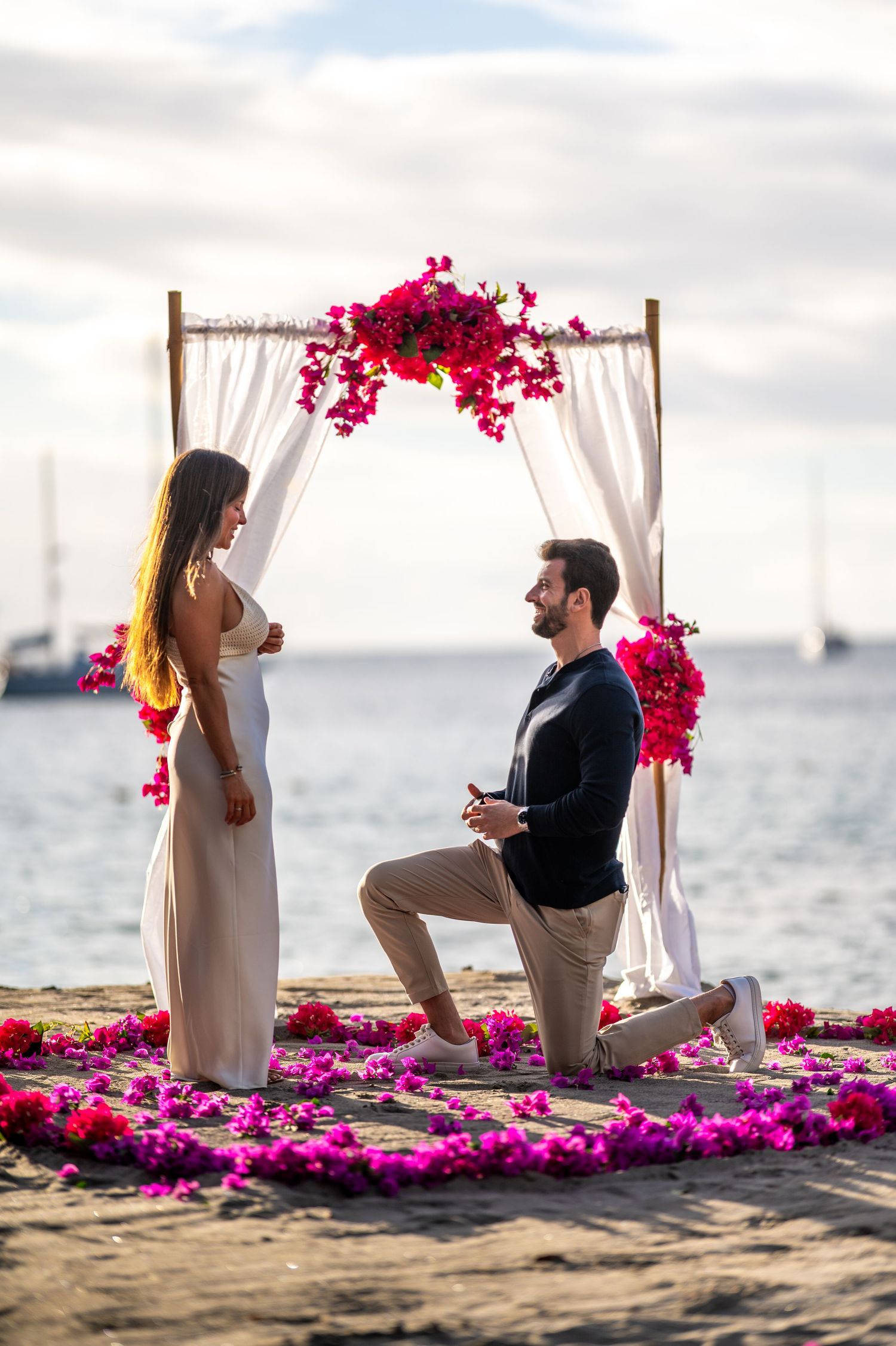 A romantic beach proposal scene with pink flowers and white curtains framing the couple at sunset.