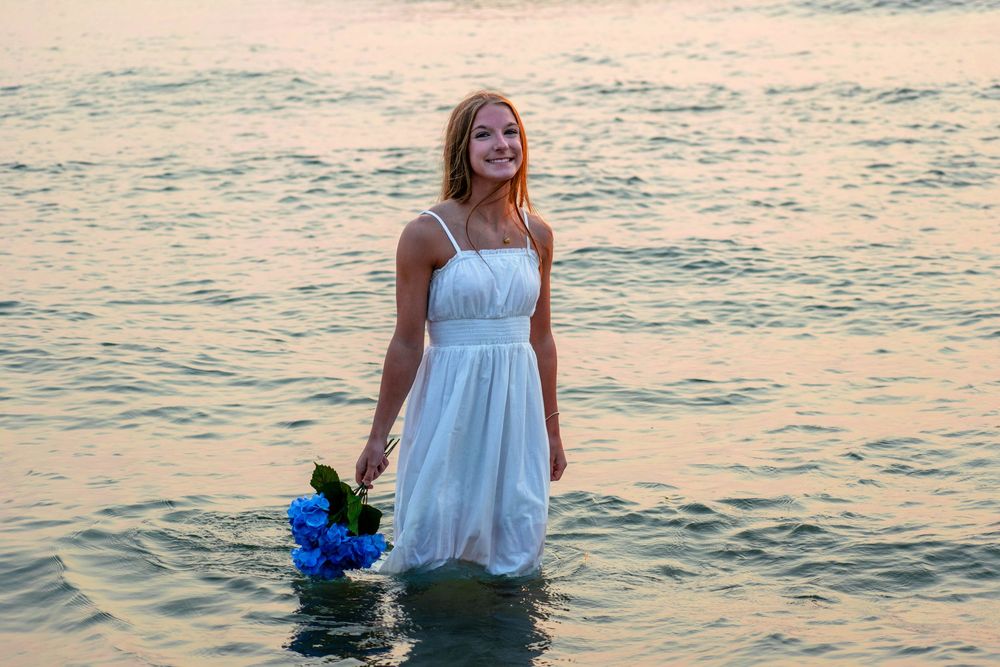 Woman in white dress poses near water during golden hour sunset.