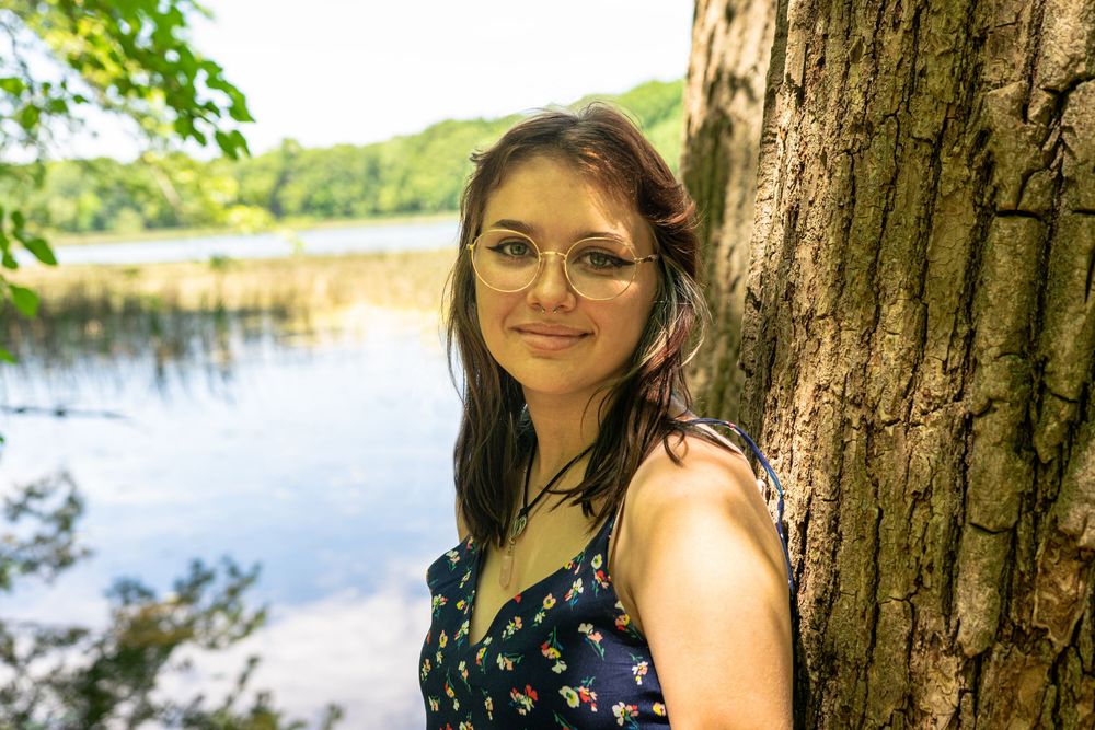 Person posing by tree trunk near lake with natural lighting.