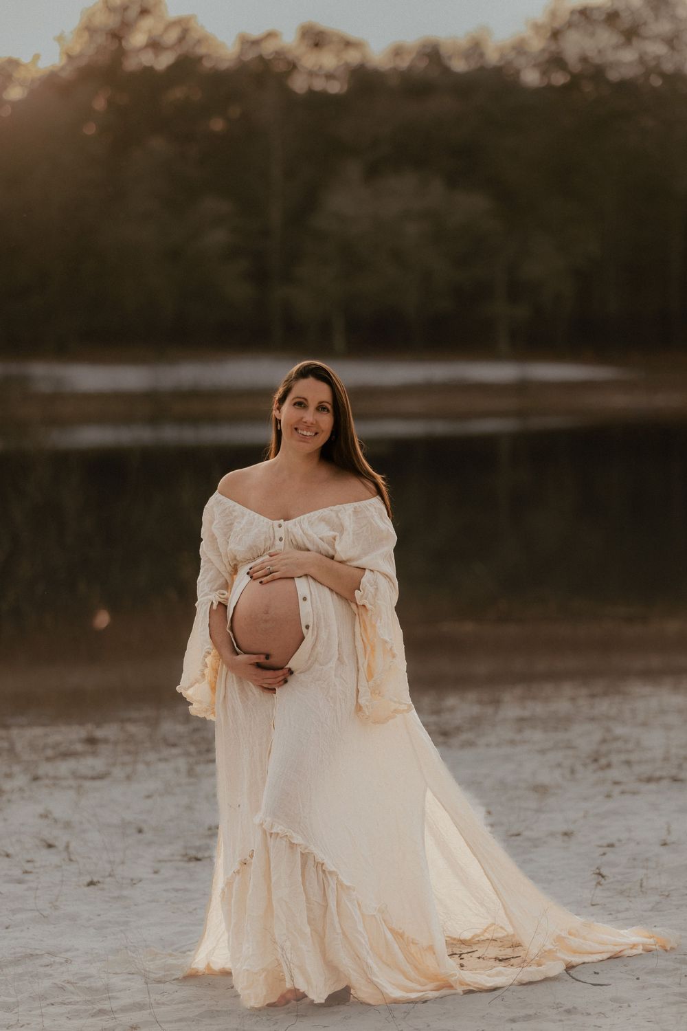 Pregnant woman in off-shoulder white boho gown cradling baby bump at golden hour near a serene lake.