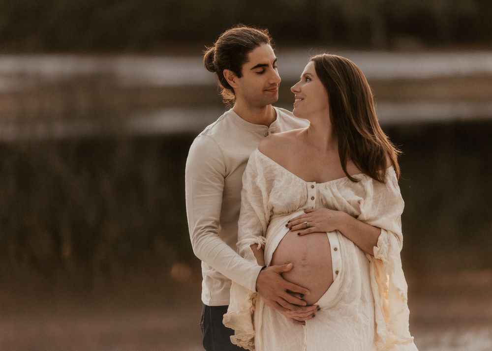 A couple shares a tender moment outdoors, the woman in a white dress showing her baby bump during a maternity photoshoot.