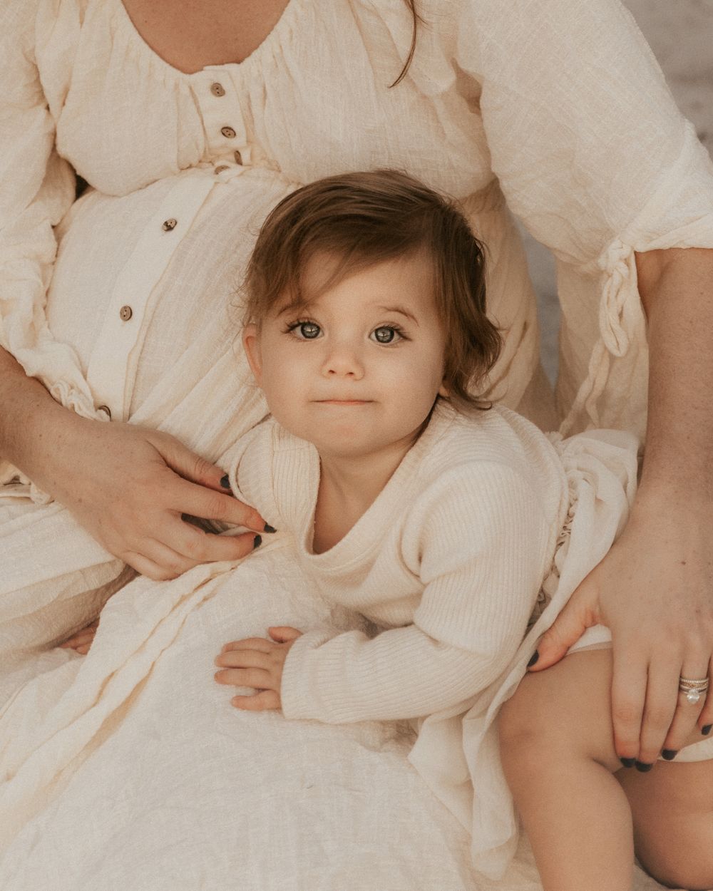 Adorable toddler in cream dress sits with pregnant mother during a soft, neutral-toned maternity photo session.
