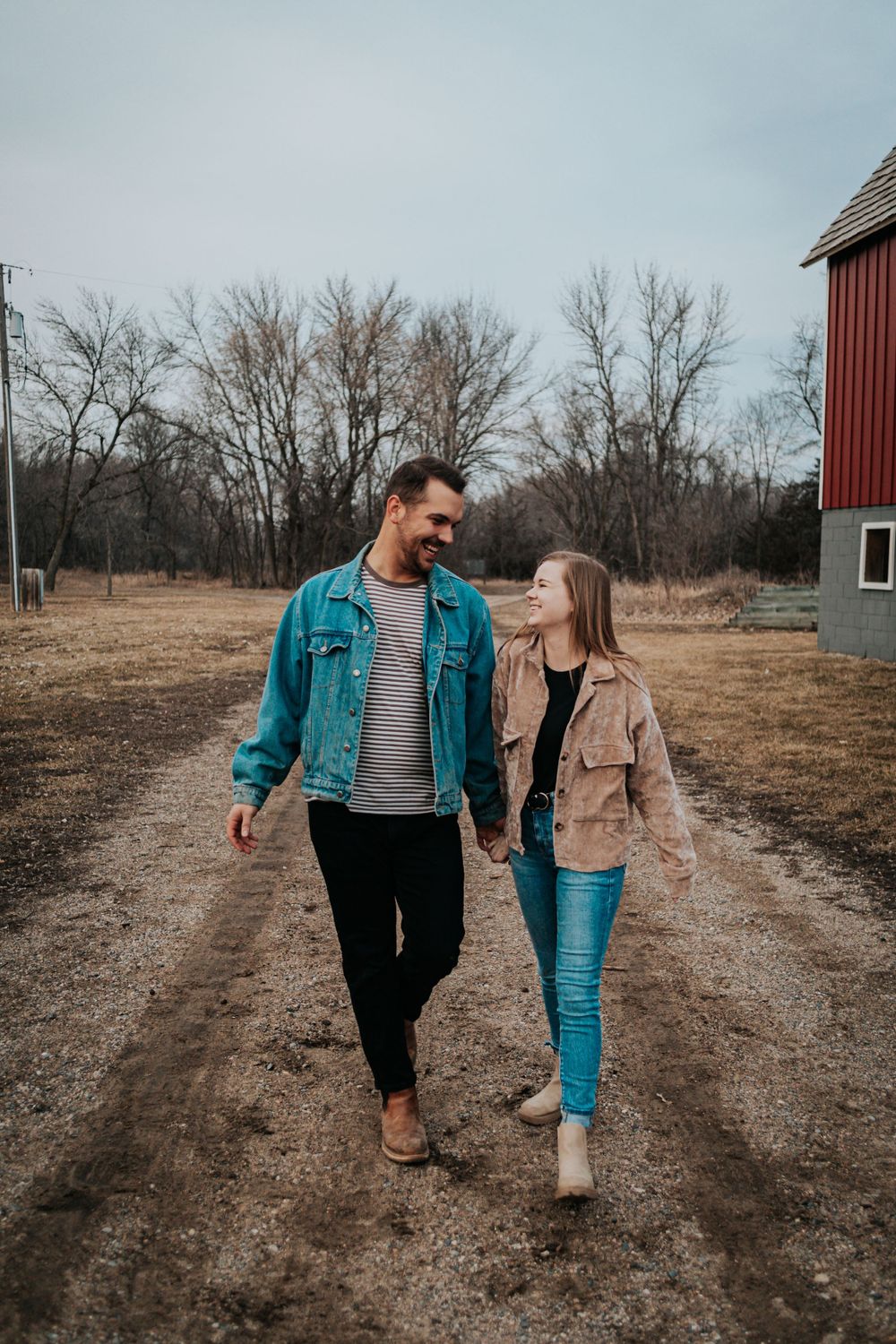 Couple walking together on a dirt path near a red barn on a cloudy autumn day.