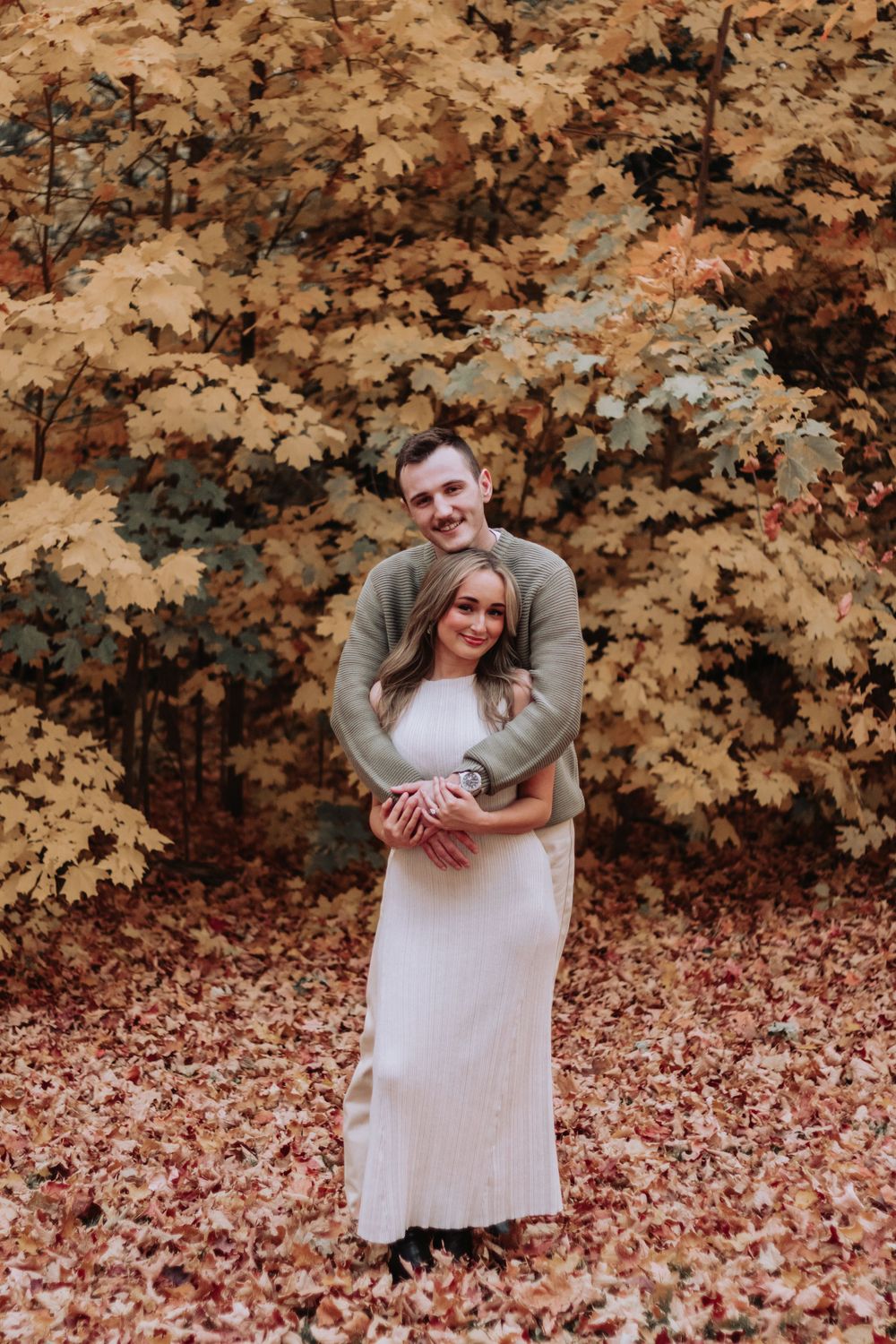 Couple embracing under golden autumn trees with fallen leaves covering the ground.