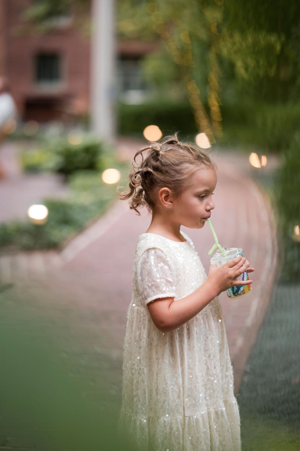 A young child in a white lace dress sips from a straw while standing outdoors in soft evening lighting.