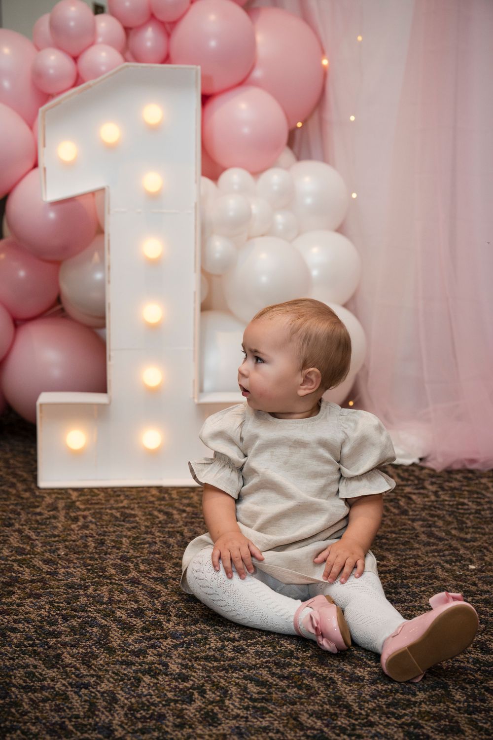 Baby in white outfit sits on patterned carpet beside illuminated number one decoration with pink balloon backdrop.