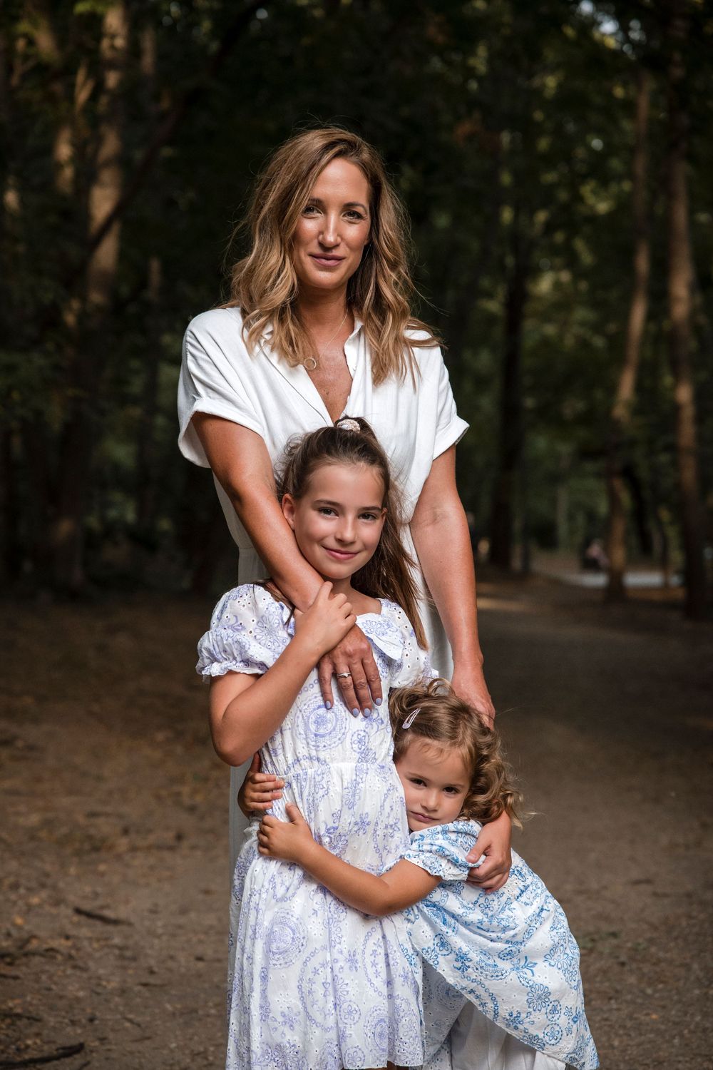 A family wearing white and blue floral dresses poses together in a wooded outdoor setting during a photo session.