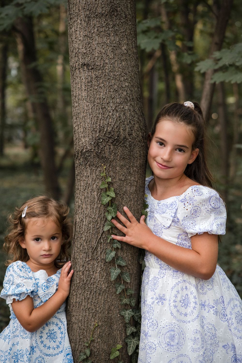 A series of photos showing two children in matching blue and white dresses posing playfully against a tree trunk in a forest.