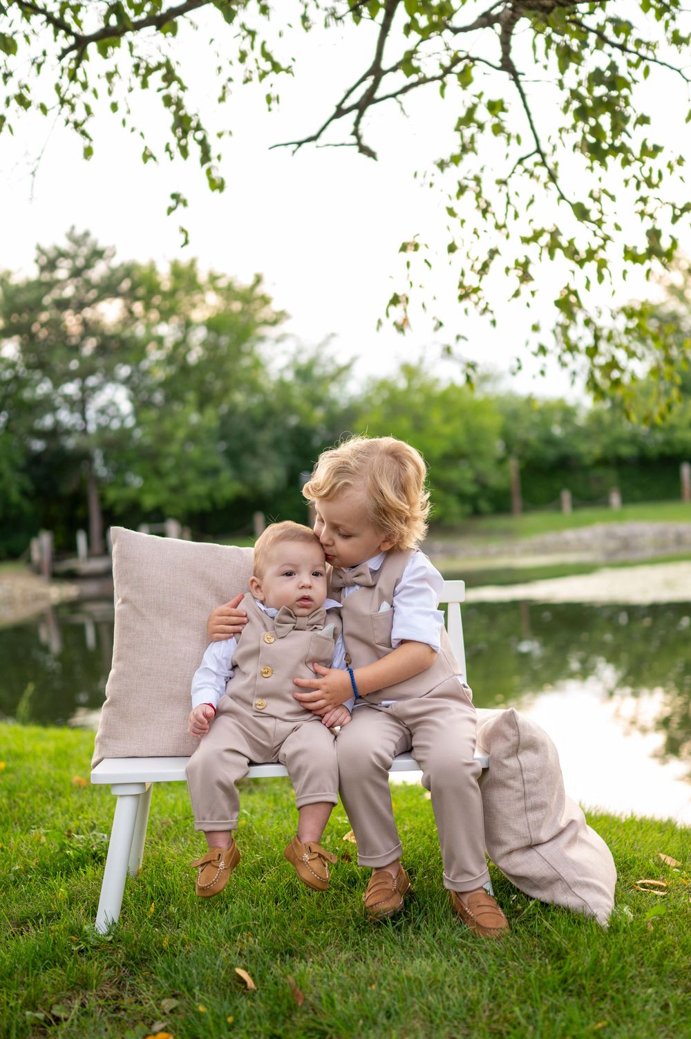 Two young children in matching beige outfits sit together on grass near a pond on a sunny day.