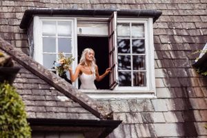 Person in white dress gazes dreamily from rustic dormer window with slate roof tiles and window flowers.
