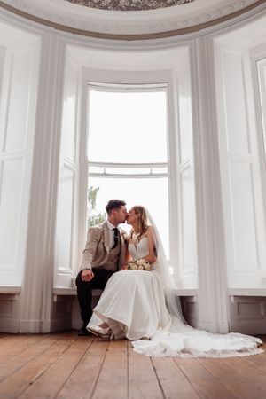 A romantic wedding moment captured in a bright bay window alcove with white walls and wood flooring.