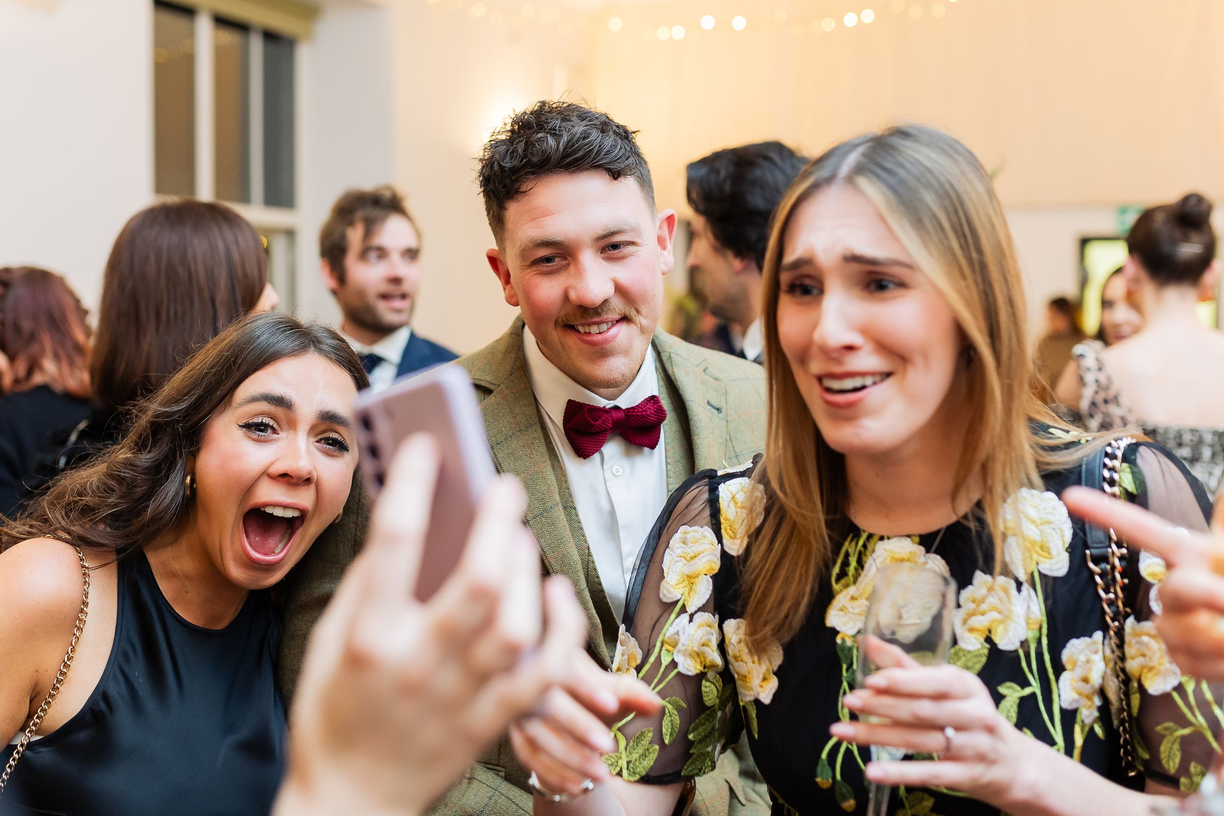 A group of wedding guests laugh and smile while holding up their phones to take photos at a celebration.
