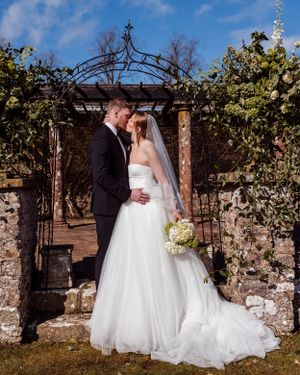 A couple shares a tender moment under a rustic garden archway with stone walls and climbing vines on a sunny day.
