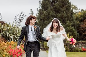 Joyful bride and groom holding hands walking through a lush garden, bride in white gown with veil holding pink bouquet.