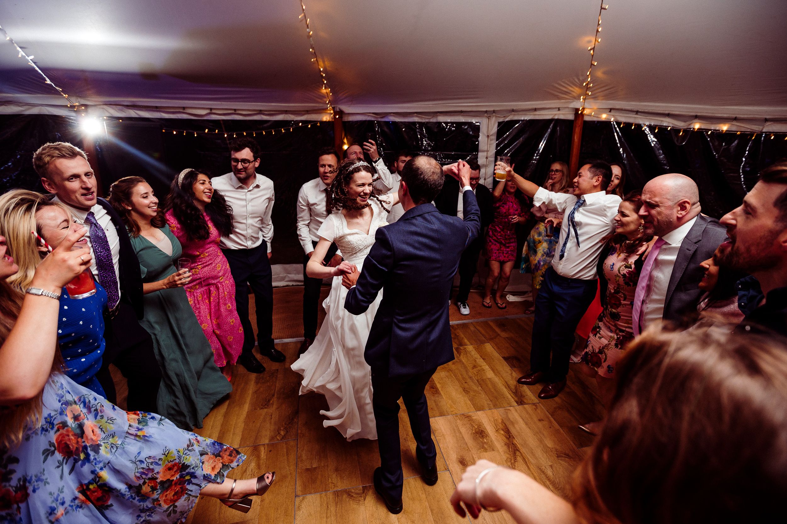 People dance and celebrate on a dance floor at a wedding reception while colored lights illuminate the scene.