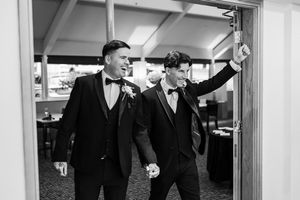 Two grooms in black tuxedos and bow ties hold hands while entering a wedding reception venue.