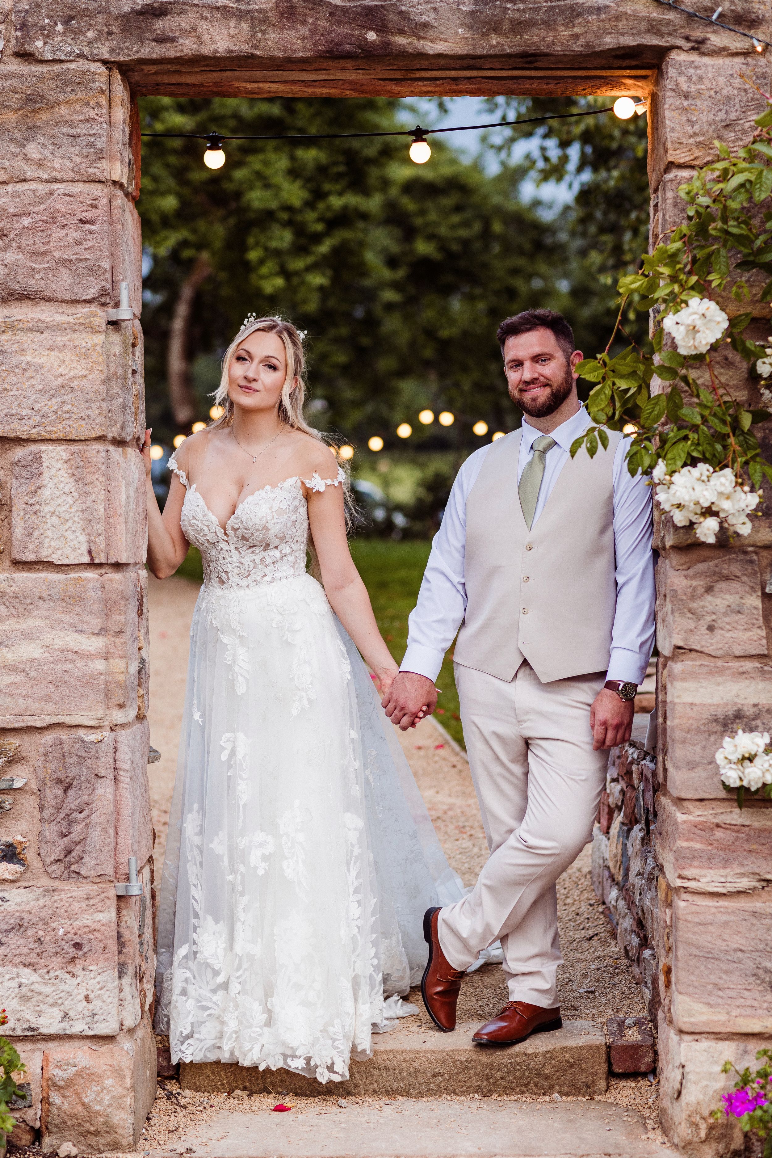 A wedding couple poses in a rustic stone doorway decorated with white flowers and string lights in a garden setting.
