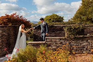Bride in a flowing white gown and veil holds hands with a groom in a gray suit on a stone wall terrace surrounded by autumn foliage.