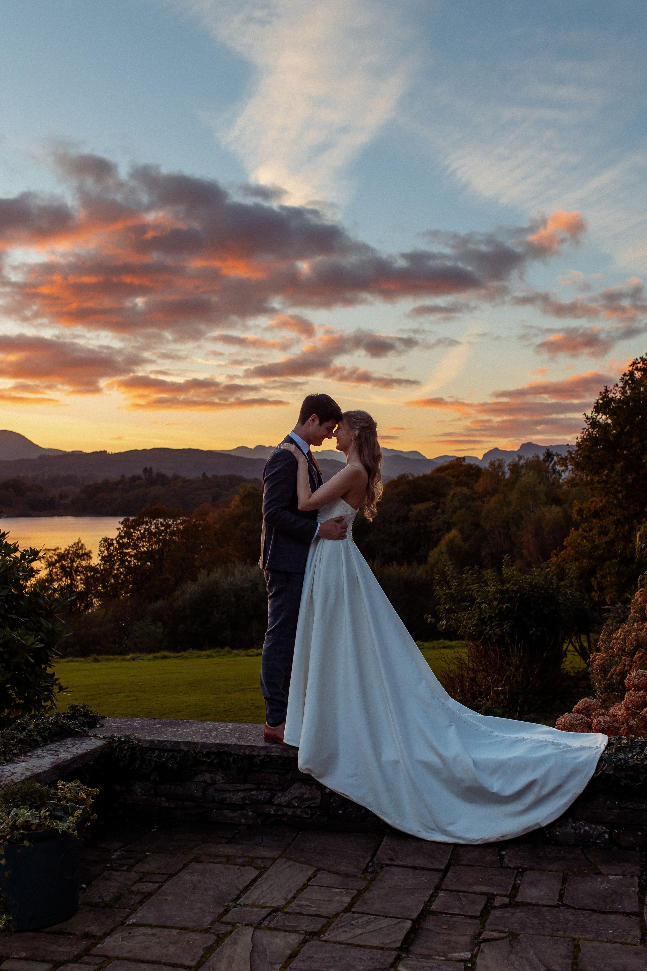 Bride in a flowing white gown and groom in a dark suit embrace on a stone terrace at sunset, with a lake and mountains behind.