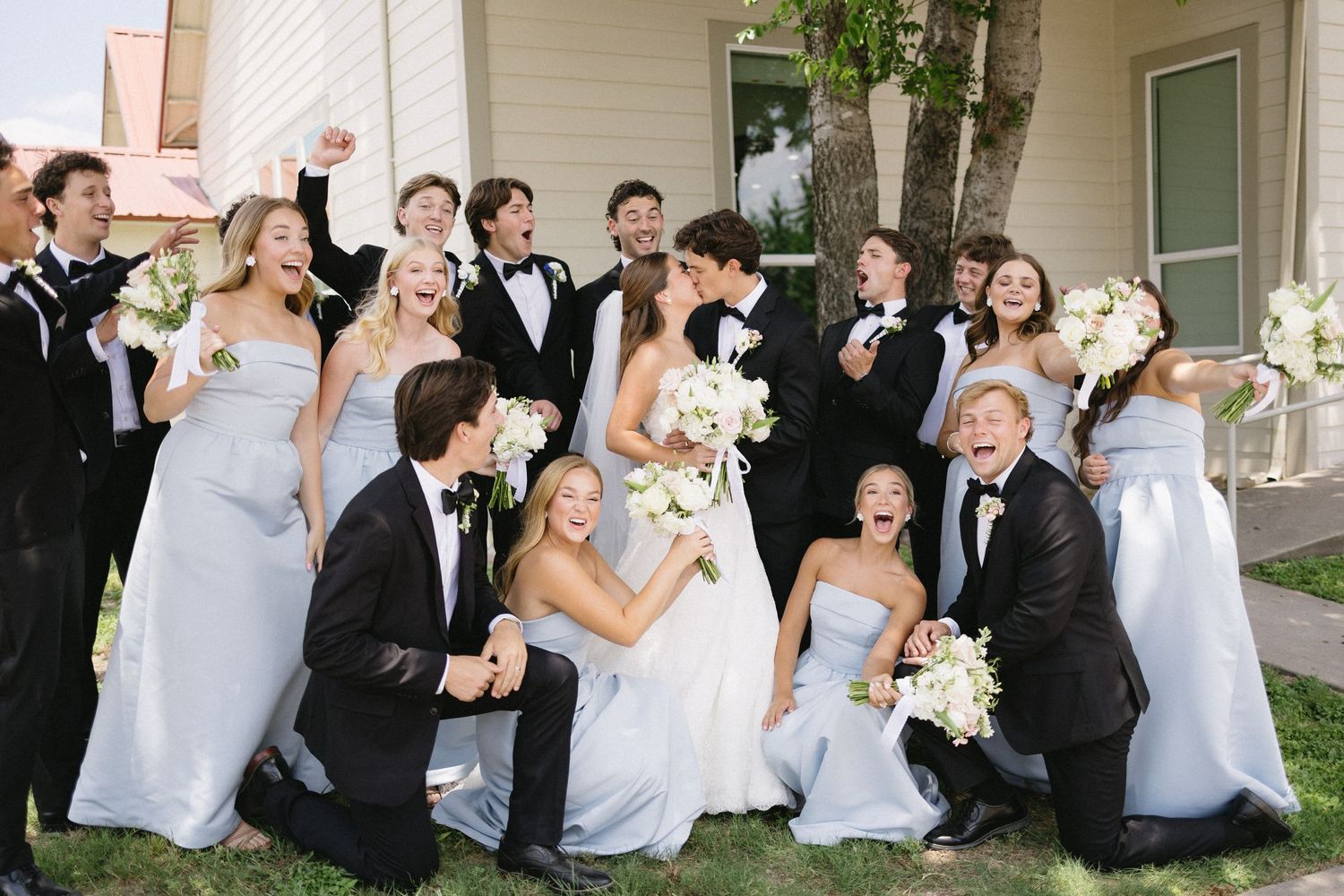 Joyful wedding party celebrating outdoors in front of a white building with bouquets and tuxedos.