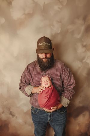 A man in plaid shirt and jeans holds a baby wrapped in red in a professional studio setting.