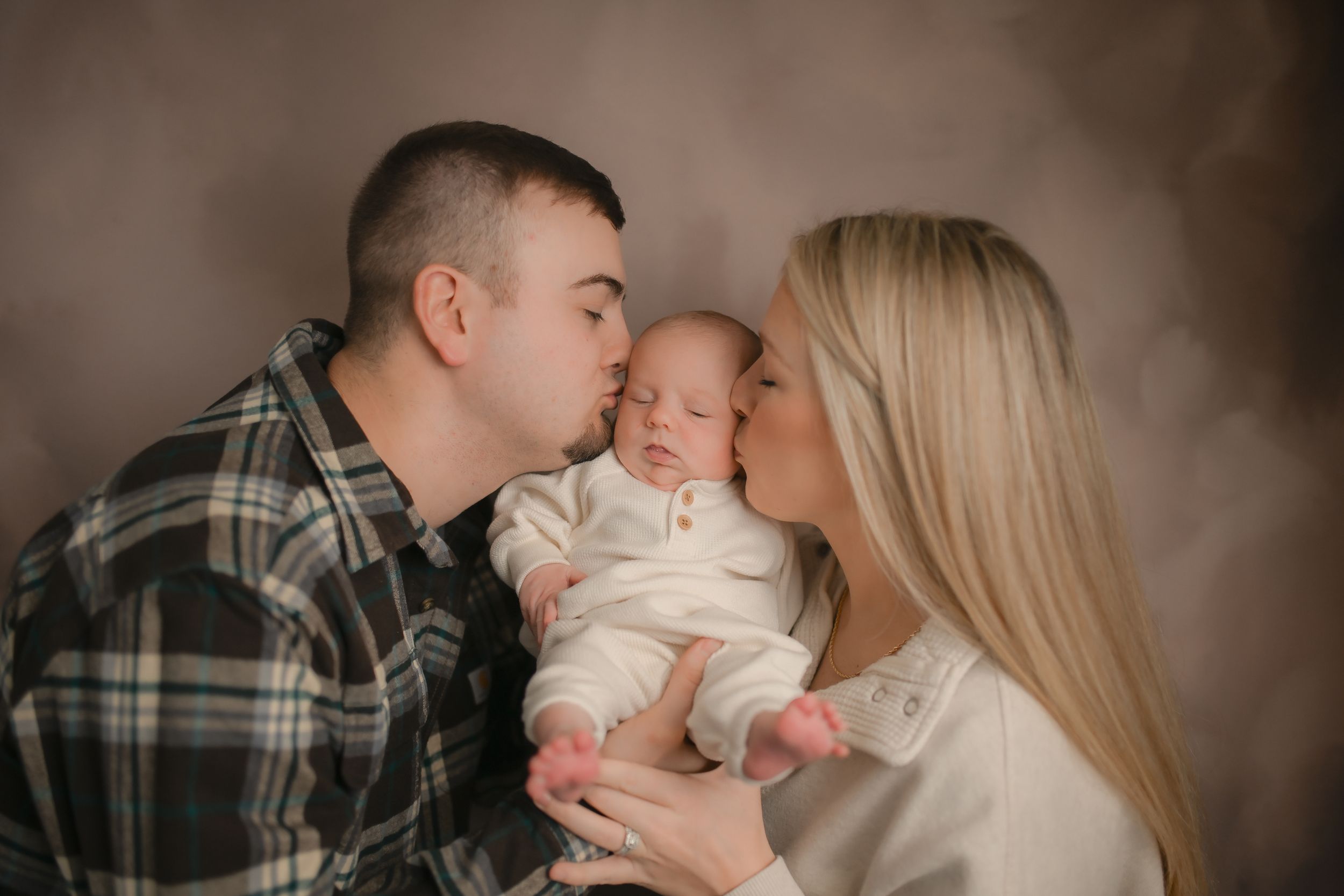 A tender family moment as parents share a kiss with their sleeping newborn baby in a studio portrait.