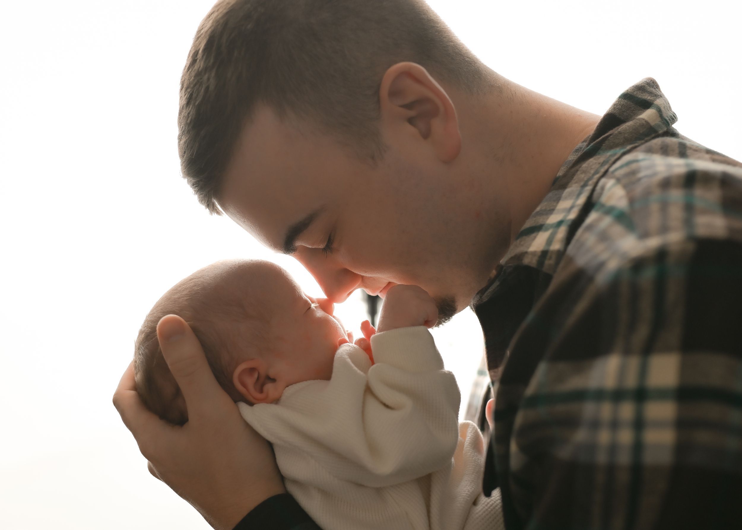 Parent sharing intimate nose-to-nose moment with baby in plaid shirt against bright window light.