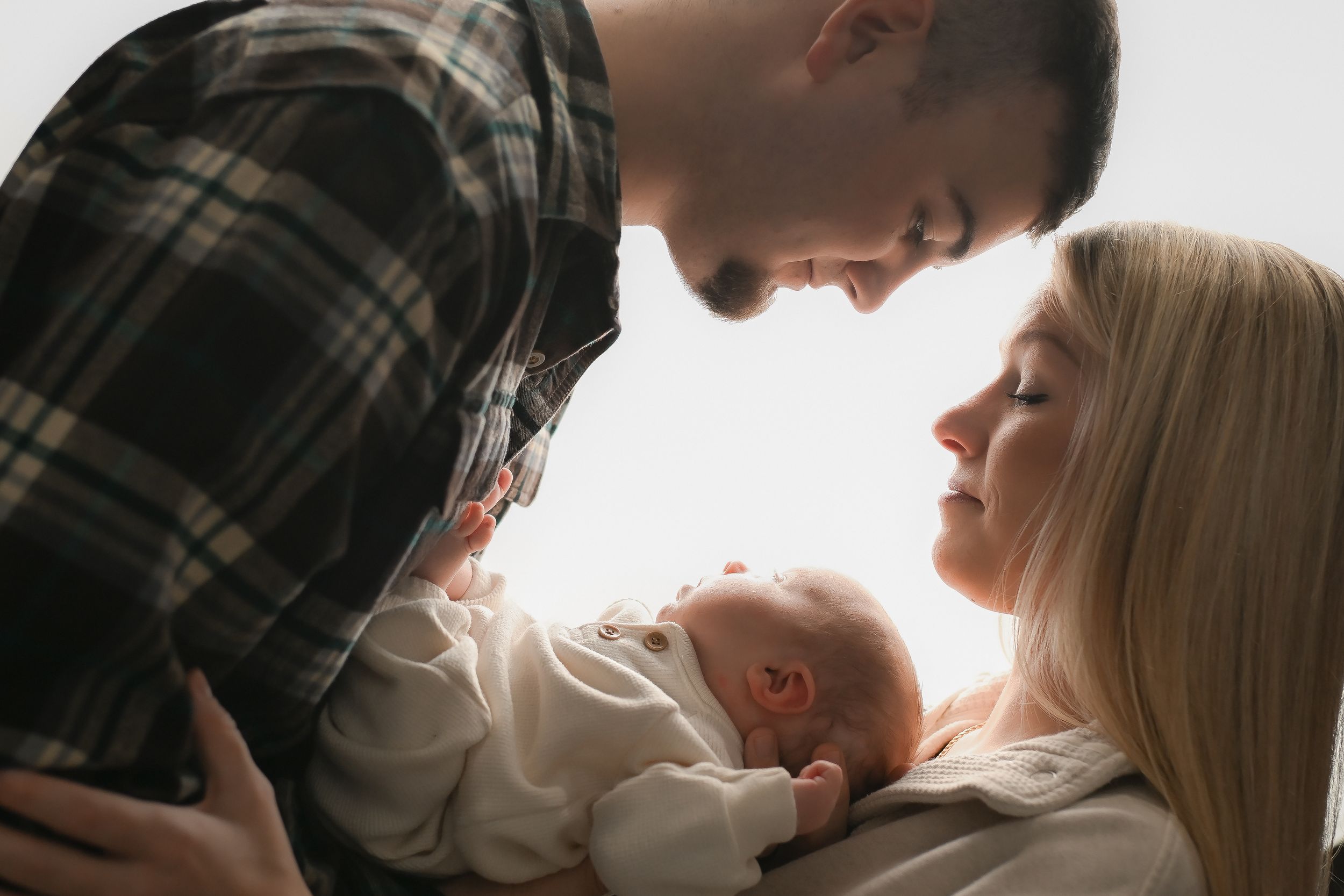Intimate family portrait showing parents lovingly gazing at their newborn baby against white background.